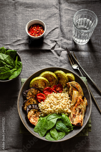 Healthy vegetarian breakfast idea. A bowl with baked vegetables, zucchini, eggplant, tomatoes, fennel, bulgur, fresh spinach. Red hot chili flakes, a glass of water, knife and fork.