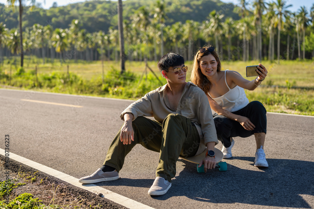 Obraz premium Young Asian Couple Lover using smartphone selfie themself while playing Surfskate on a Road, Summer Season