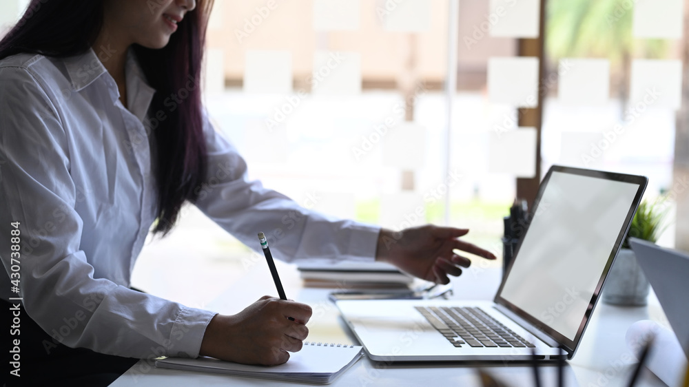 Cropped shot of young female office worker making note on notebook and pointing on screen of laptop computer. Cropped shot of young female office worker making note on notebook and pointing on screen of laptop computer.