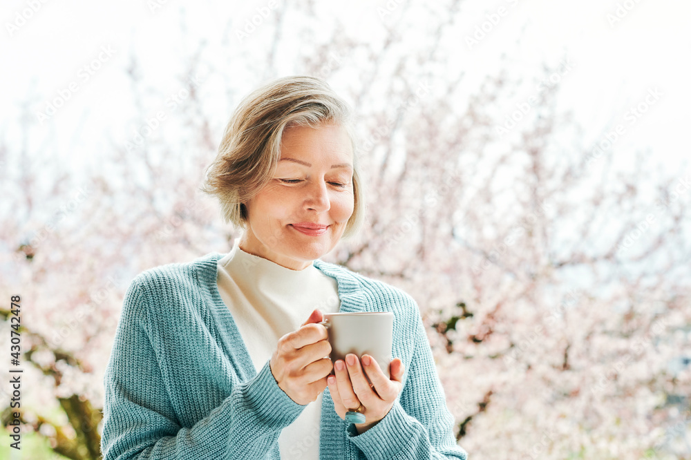Spring portrait of beautiful and elegant middle age woman posing against pink blooming tree, holding cup of coffee or tea Spring portrait of beautiful and elegant middle age woman posing against pink blooming tree, holding cup of coffee or tea