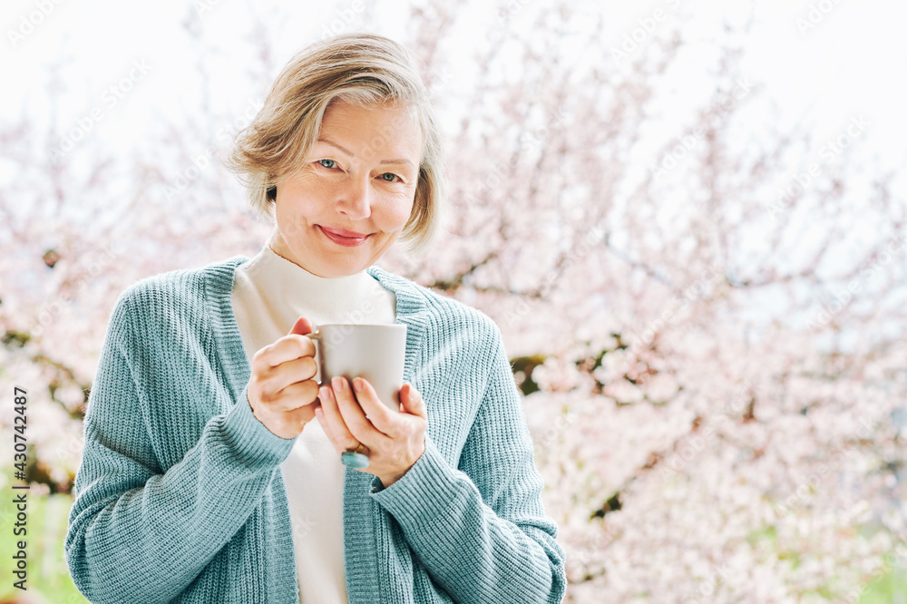 Spring portrait of beautiful and elegant middle age woman posing against pink blooming tree, holding cup of coffee or tea Spring portrait of beautiful and elegant middle age woman posing against pink blooming tree, holding cup of coffee or tea