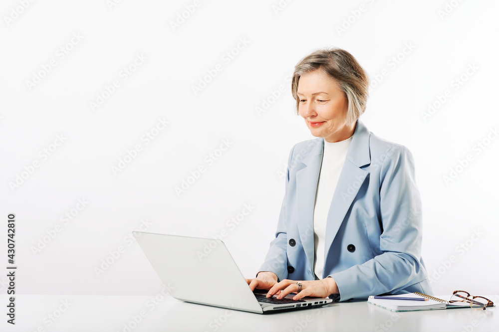 Studio portrait of middle age woman posing on white background, sitting at the desk, using laptop Studio portrait of middle age woman posing on white background, sitting at the desk, using laptop