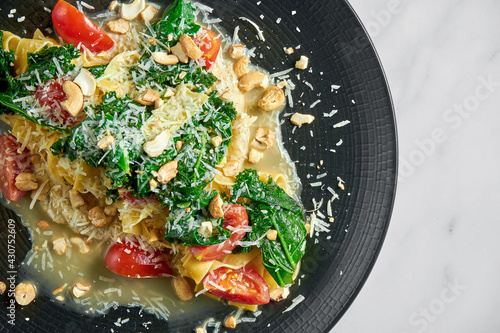 Pappardelle pasta with parsnip, cream sauce, blanched greens, tomatoes and nuts on a black plate on a white marble background