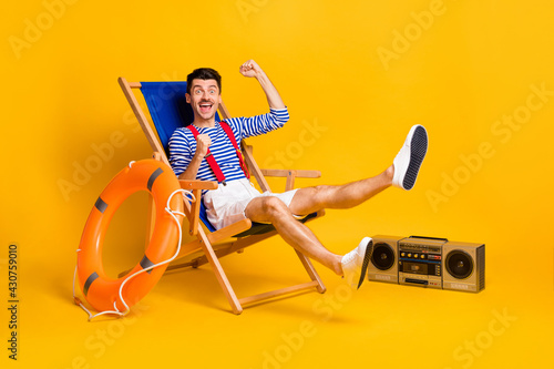 Full body photo of happy victorious young man sit chair beach raise fists isolated on yellow color background