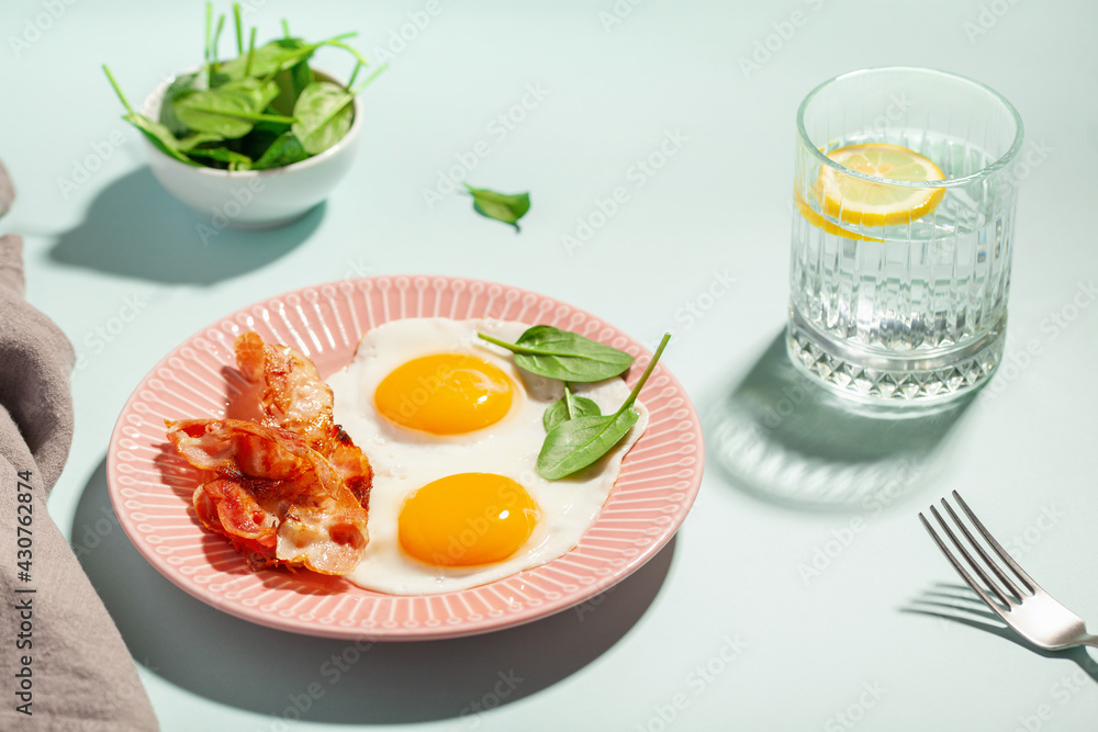 English breakfast - eggs, bacon and spinach leaves in a plate. Water with lemon. Hard light. English breakfast - eggs, bacon and spinach leaves in a plate. Water with lemon. Hard light.