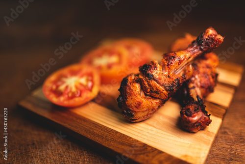 Close of roasted chicken drumsticks decorated on wooden plate along with fresh, juicy tomatoes. Dramatic image of non vegetarian food with copy space for text.