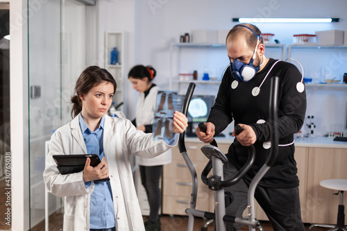 Medical doctors in sports science lab measuring performance cardiorespiratory of athlete. Team of researcher monitoring vo2 of sportsman running on cross trainer equipped with electrodes and mask. photo