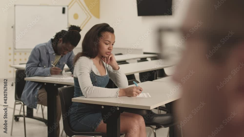 Medium long of diverse multiethnic students sitting at desks in classroom, writing on exam, then camera focusing on unrecognizable teacher wearing glasses, watching them Medium long of diverse multiethnic students sitting at desks in classroom, writing on exam, then camera focusing on unrecognizable teacher wearing glasses, watching them