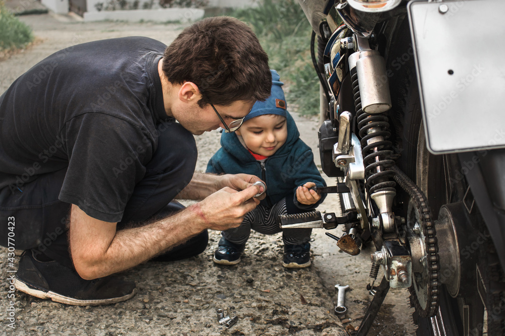 In the afternoon, a father and son repair a black motorcycle on the street. Communication between father and son.