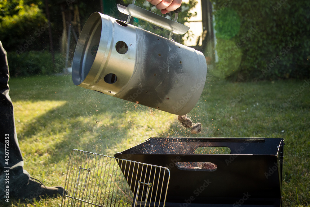 Mann gibt glühende Kohle von Anzündkamin in Klappgrill nach Anheizen für Glut zum grillen draußen im Garten Mann gibt glühende Kohle von Anzündkamin in Klappgrill nach Anheizen für Glut zum grillen draußen im Garten