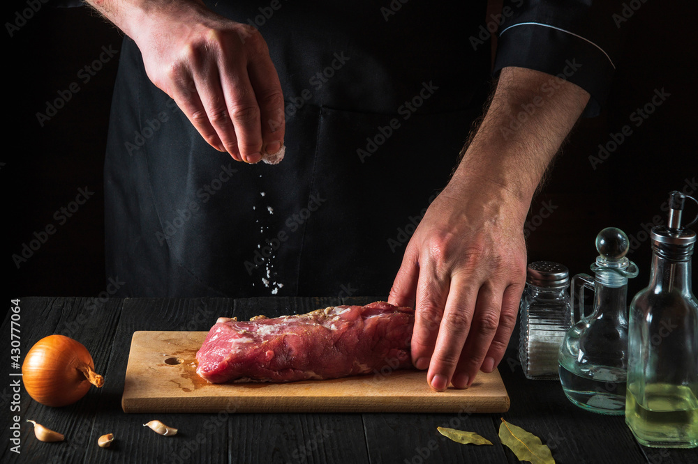 Chef or cook sprinkles the raw beef meat with salt. Preparing meat before baking. Working environment in kitchen of restaurant or cafe