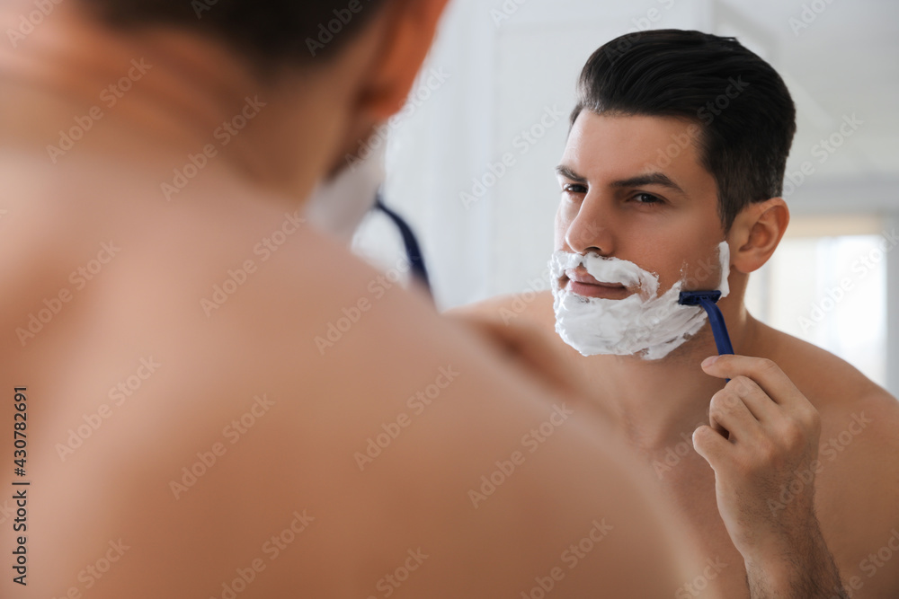 Handsome man shaving near mirror in bathroom