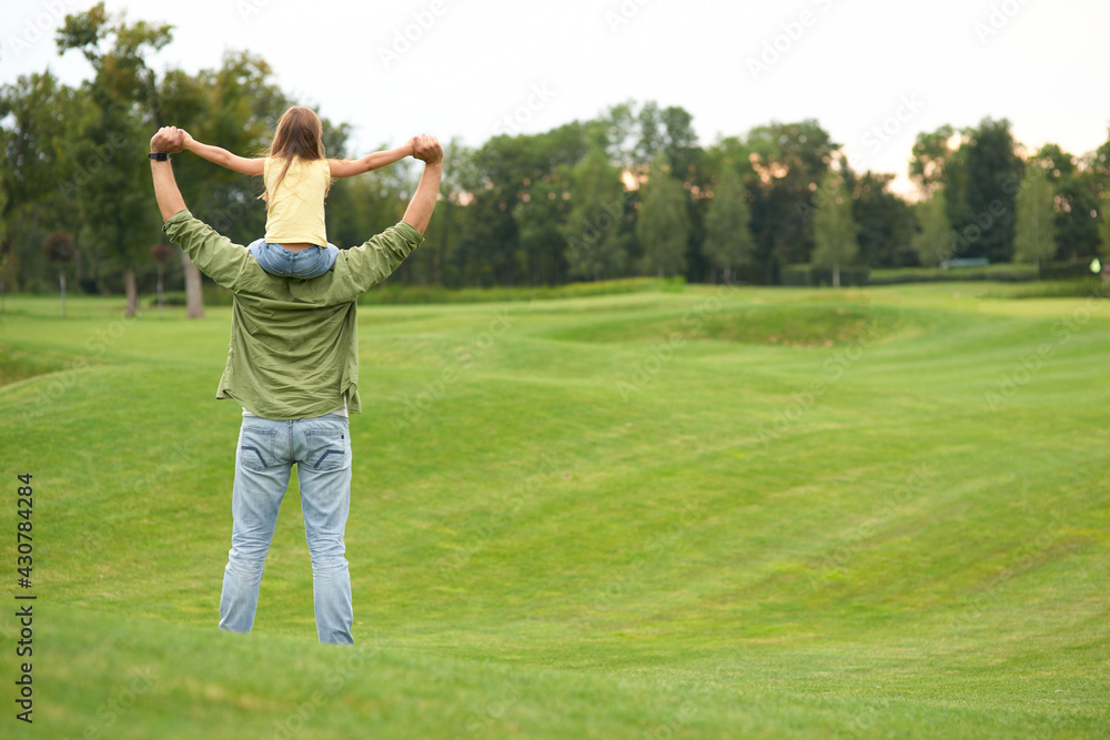 Rear view of little girl sitting on father's shoulders and holding each other hands, standing in a green field on a warm day, spending time together outdoors