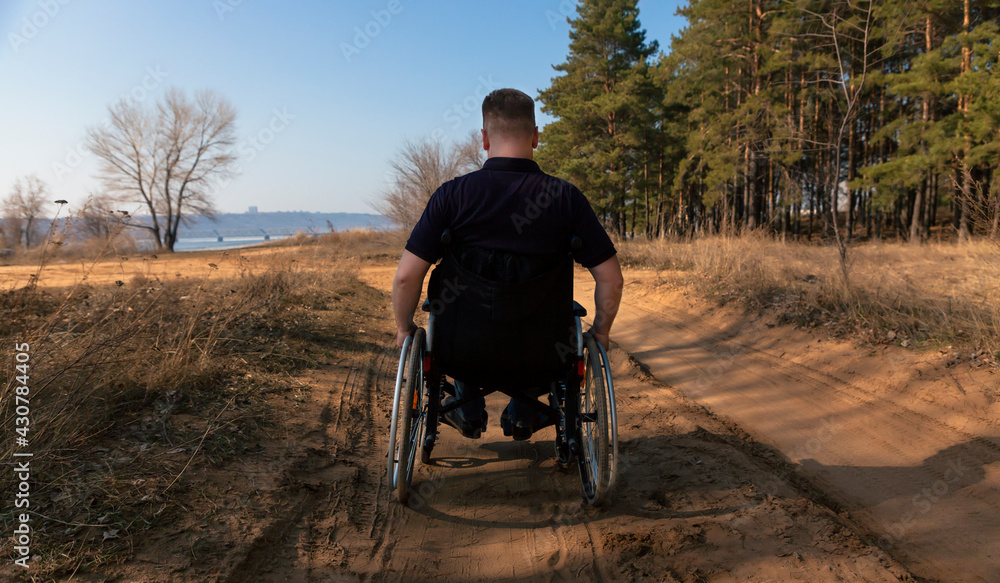 A man rides in a wheelchair on the road. A disabled man in a wheelchair rides heavily on a sandy road along the forest. Rear view. Selective focus A man rides in a wheelchair on the road. A disabled man in a wheelchair rides heavily on a sandy road along the forest. Rear view. Selective focus
