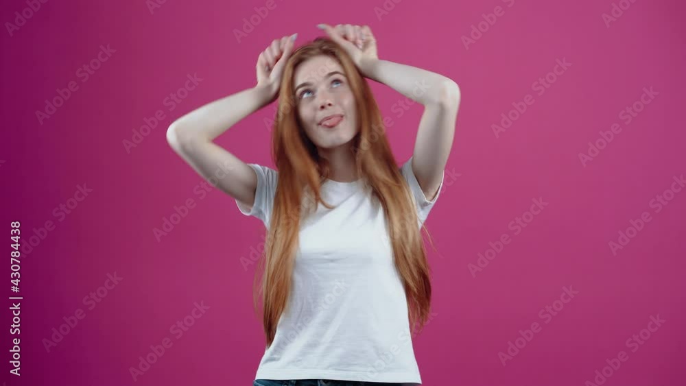 The portrait of a charismatic young woman who playfully puts her hands to her head like bunny ears. Freckled teenage girl in a white T-shirt, isolated on a pink background. The concept of people's The portrait of a charismatic young woman who playfully puts her hands to her head like bunny ears. Freckled teenage girl in a white T-shirt, isolated on a pink background. The concept of people's
