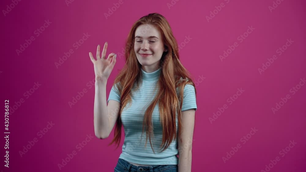 The portrait of a beautiful, cheerful red-haired girl shows the ok sign and blinks with chic eyes.Freckled teenage girl in a blue T-shirt, isolated on a pink background. The concept of people's The portrait of a beautiful, cheerful red-haired girl shows the ok sign and blinks with chic eyes.Freckled teenage girl in a blue T-shirt, isolated on a pink background. The concept of people's