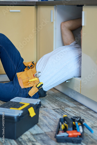 Get fixed. Professional plumber wearing tool belt examining and fixing sink pipe indoors. Tools and equipment on the floor in the kitchen