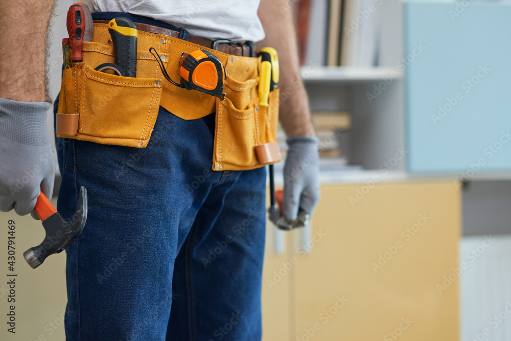 Cropped shot of professional repairman wearing a tool belt, holding a hammer and an adjustable wrench in his hands while standing indoors Cropped shot of professional repairman wearing a tool belt, holding a hammer and an adjustable wrench in his hands while standing indoors