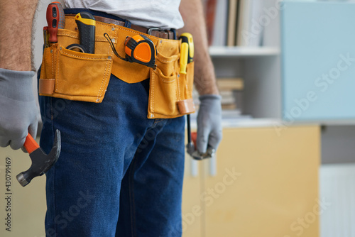Cropped shot of professional repairman wearing a tool belt, holding a hammer and an adjustable wrench in his hands while standing indoors © Kostiantyn