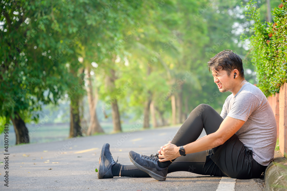 A young Asian runner sits down on a park road due to severe pain in his ankle. The concept of exercise injuries. A young Asian runner sits down on a park road due to severe pain in his ankle. The concept of exercise injuries.