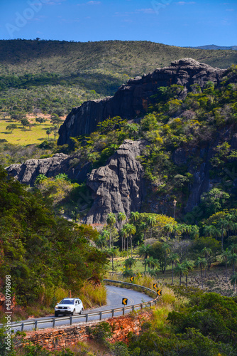 A passenger vehicle rounding a bend on a mountain road through the lush and rocky cerrado landscape of Serra do Cipo National Park, Minas Gerais, Brazil