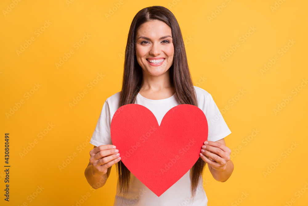Photo portrait of happy smiling woman demonstrating red paper heart postcard isolated on vibrant yellow color background Photo portrait of happy smiling woman demonstrating red paper heart postcard isolated on vibrant yellow color background