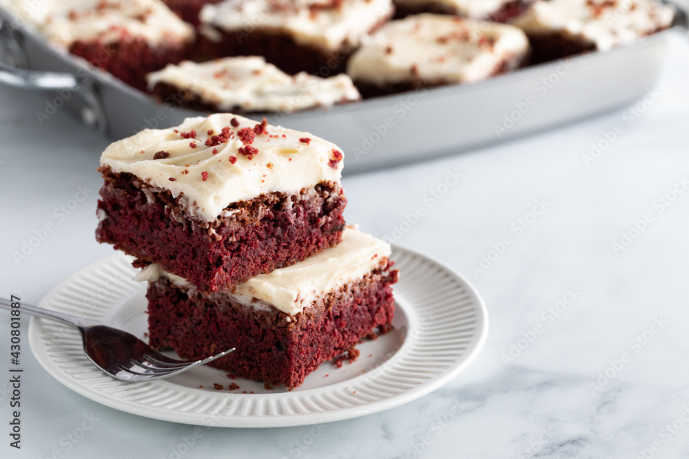 Two red velvet squares in a stack on a plate with other squares in soft focus in behind and copy space to the right. Two red velvet squares in a stack on a plate with other squares in soft focus in behind and copy space to the right.