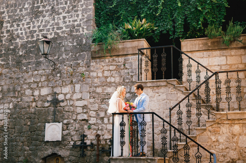 The bride and groom stand on a beautiful staircase with a wrought iron railing in the old town of Perast and look at each other  © Nadtochiy