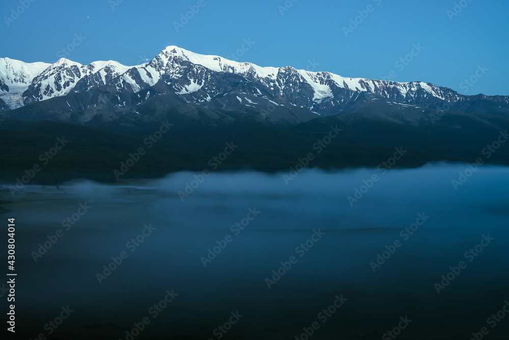 Atmospheric landscape with dense fog on lake and great snowy mountain ridge under night sky. Alpine scenery with thick fog on mountain lake and big mountain range in night. Snow pinnacle in dusk.