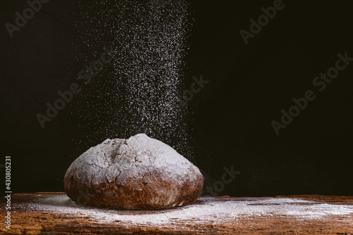 rustic and golden round loaf of fresh whole grain bread on dark black background on top of wooden kitchen table with cascade of falling flour