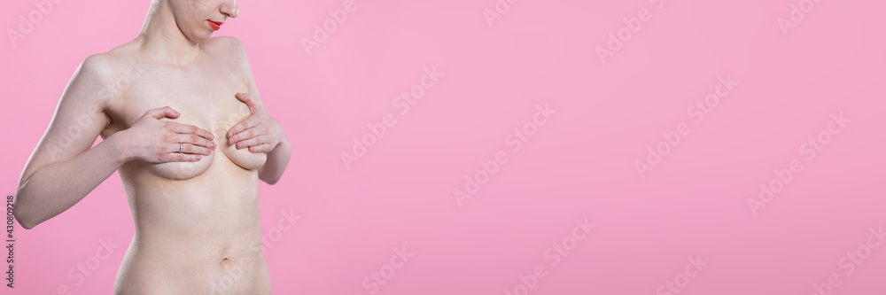 Young nude woman gently covers her breasts with both hands. Portrait on pink background. Panoramic frame. Young nude woman gently covers her breasts with both hands. Portrait on pink background. Panoramic frame.