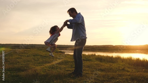 Little daughter plays with dad in the park in the sun. Dad shakes the hands of a happy child in the rays of the sunset in the park. Happy family on vacation. A cheerful kid plays with his father. © zoteva87