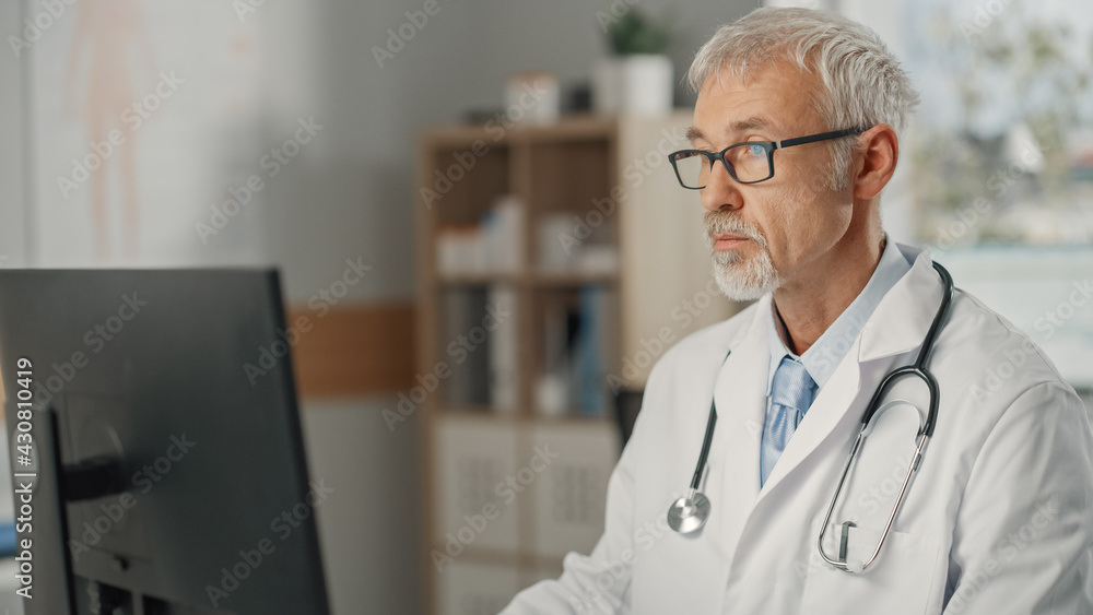 Experienced Middle Aged Male Doctor Wearing White Coat Working on Personal Computer at His Office. Senior Medical Health Care Professional Working with Test Results, Patient Treatment Planning. Experienced Middle Aged Male Doctor Wearing White Coat Working on Personal Computer at His Office. Senior Medical Health Care Professional Working with Test Results, Patient Treatment Planning.