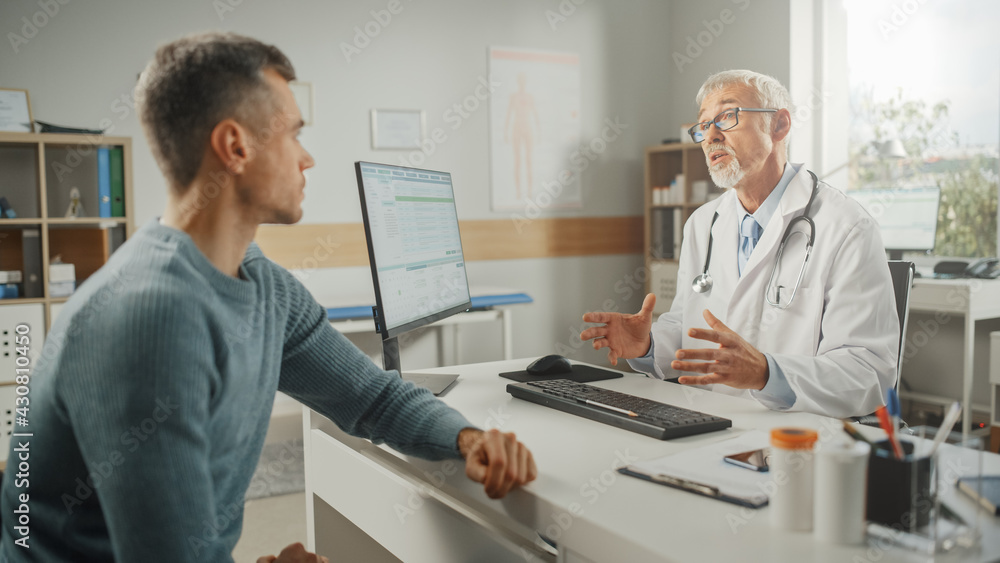 Middle Aged Family Doctor is Talking with Young Male Patient During Consultation in a Health Clinic. Senior Physician in Lab Coat Sitting Behind a Computer Desk in Hospital Office. Middle Aged Family Doctor is Talking with Young Male Patient During Consultation in a Health Clinic. Senior Physician in Lab Coat Sitting Behind a Computer Desk in Hospital Office.