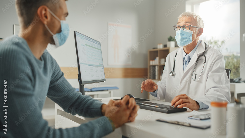 Middle Aged Family Doctor is Talking with Young Male Patient During Consultation in a Health Clinic. Both Wear Face Masks. Physician in Lab Coat Sitting Behind a Computer Desk in Hospital Office. Middle Aged Family Doctor is Talking with Young Male Patient During Consultation in a Health Clinic. Both Wear Face Masks. Physician in Lab Coat Sitting Behind a Computer Desk in Hospital Office.