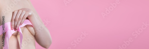 A young and naked woman holds a pink ribbon with one hand and covers her breasts. A symbol of the fight against breast cancer. Panoramic frame.