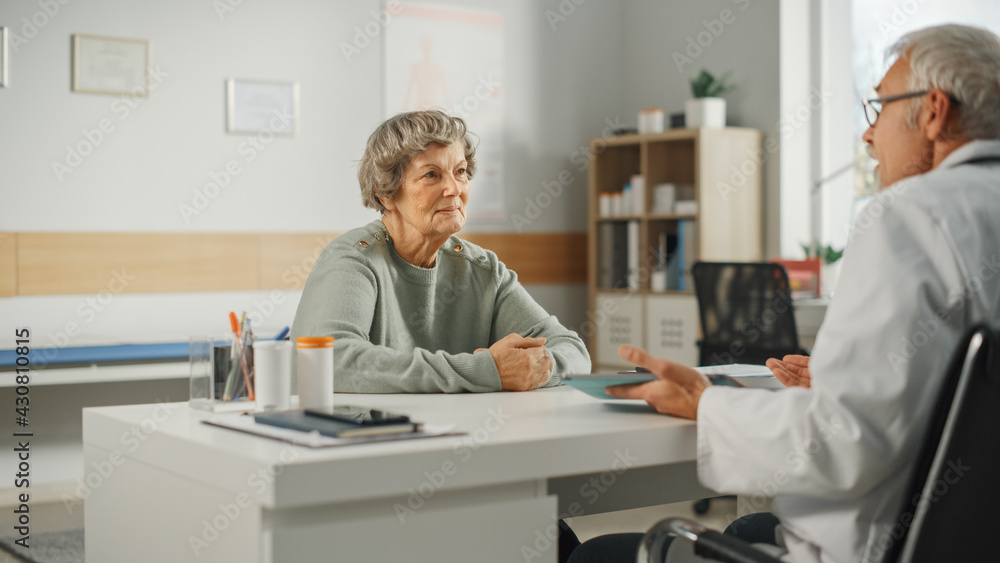 Middle Aged Family Doctor is Talking with Senior Female Patient During Consultation in a Health Clinic. Experienced Caucasian Physician in Lab Coat Sitting Behind a Computer Desk in Hospital Office.
