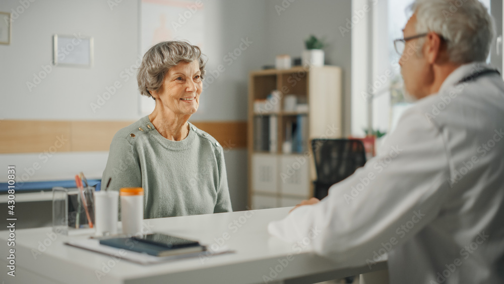 Obraz premium Middle Aged Family Doctor is Talking with Senior Female Patient During Consultation in a Health Clinic. Experienced Caucasian Physician in Lab Coat Sitting Behind a Computer Desk in Hospital Office.