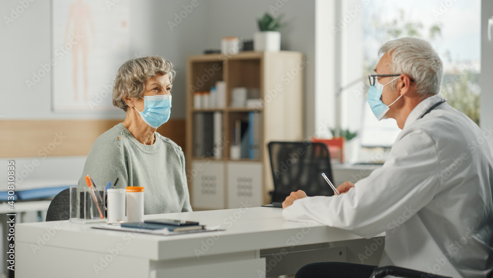 Obraz premium Middle Aged Family Doctor is Talking with Senior Female Patient During Consultation in a Health Clinic. Both are Wearing Face Masks. Physician in Lab Coat Sitting Behind a Desk in Hospital Office.