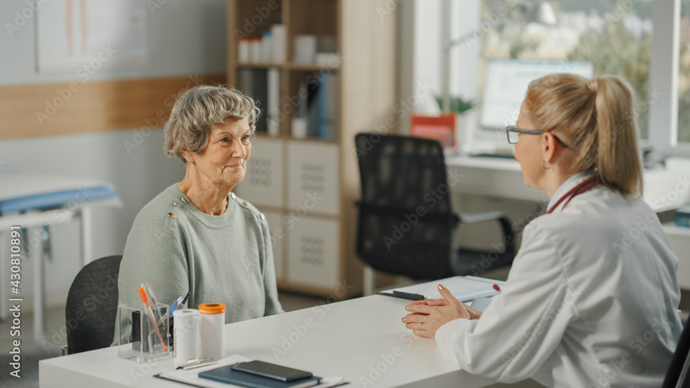 Fototapeta premium Female Family Doctor is Sitting Behind a Desk and Speaking to a Senior Woman During Consultation in a Health Clinic. Physician in Lab Coat Prescribing Drugs to Elderly Patient in Hospital Office.