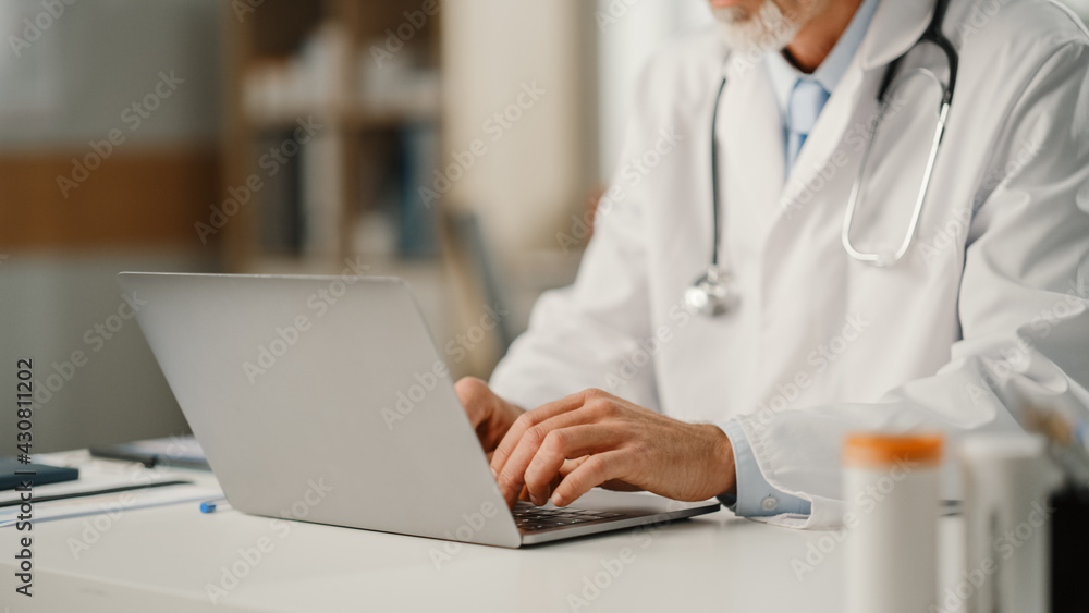 Experienced Middle Aged Male Doctor Wearing White Coat Working on Laptop Computer at His Office. Senior Medical Health Care Professional Working with Test Results, Patient Treatment Planning. Experienced Middle Aged Male Doctor Wearing White Coat Working on Laptop Computer at His Office. Senior Medical Health Care Professional Working with Test Results, Patient Treatment Planning.