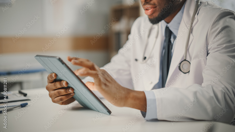 Close Up of African American Male Doctor Wearing White Coat Working on Tablet Computer at His Office. Medical Health Care Professional Working with Test Results, Patient Treatment Planning.