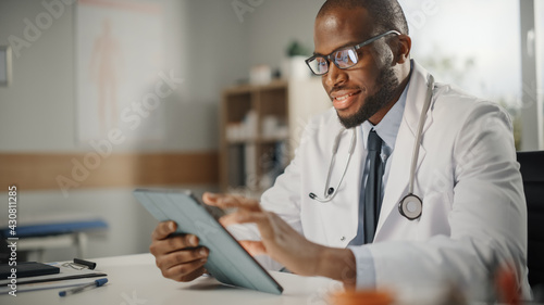 Slika na platnu Happy and Smiling African American Male Doctor Wearing White Coat Working on Tablet Computer at His Office