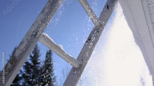POV under a ladder leaning on the edge of a roof, shooting shovelfuls of snow falling as someone on the roof throws them down. Sunny winter day