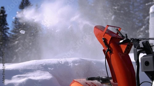 Very close up shooting of fresh snow being energetically shot in the air by the mouth of a bright orange mechanical snowplough in a sunny winter day.