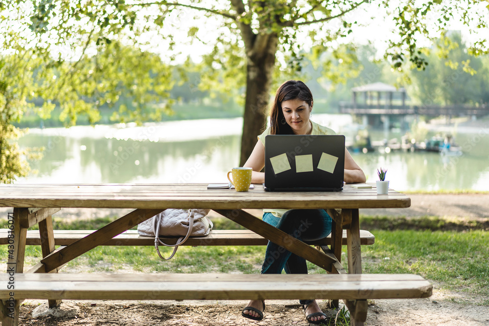 Fototapeta premium Young woman using laptop computer in the public park - Millennial female freelancer working remotely and taking notes in a city park - Technology and remote work concept - Using tech devices outdoors