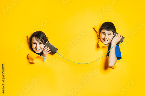 A little girl and boy playing with a handmade toy phone with a paper cup and thread. Children look into holes in torn yellow paper wall. Communication technology, information transfer concept. photo