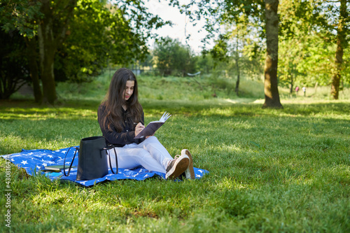 beautiful long-haired woman writing in a brown notebook in nature sitting on the grass with a blanket and her bag with trees in the background. teenage girl drawing in a notebook in the park smiling