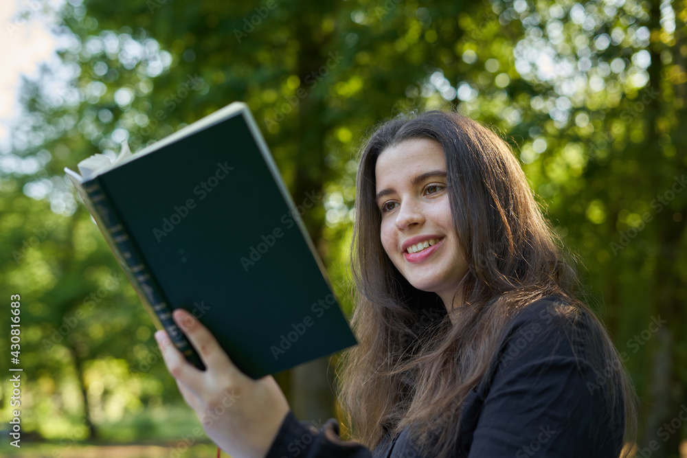 beautiful long-haired woman reading a green book in nature sitting on the grass with a blanket and her bag with trees in the background. teenager looking at a literary work in the park smiling beautiful long-haired woman reading a green book in nature sitting on the grass with a blanket and her bag with trees in the background. teenager looking at a literary work in the park smiling