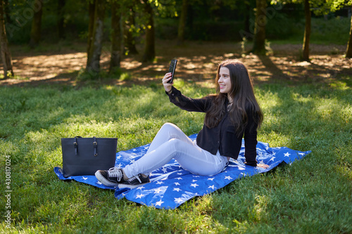 young woman with long hair in a black shirt and white pants sitting on a blanket of stars on the grass in a park with her bag using her phone to take pictures and video conferencing. influencer nature